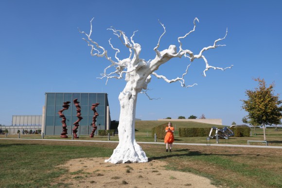 The sculpture autumn moon by artist Ugo Rondinone in front of the Museum Würth 2 in Künzelsau. It is a lifelike artificial leafless white tree stretching its gnarled branches towards the sky. In the background, more sculptures from the sculpture garden can be seen.